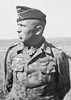 The head and shoulders of an elderly man, shown in partial profile. He wears a field cap and a military uniform and an Iron Cross displayed at the front of his camouflage shirt collar.