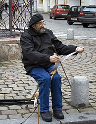 Someone on the street sitting on a bin playing a saw with a bow and with it between his legs.