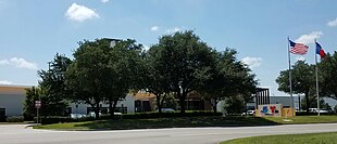 A one-story office building with K X A S and K X T X signage, mostly obscured by trees.