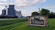 Sign at the side of a state highway reading "Welcome to Climax, established 1896". Large grain elevators are visible in the background.