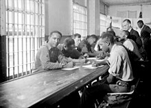 Image en noir et blanc de plusieurs prisonniers, pour la plupart d'origine africaine, assis à un bureau et écrivant. Les fenêtres sont munies de barreaux.