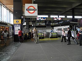 Passengers cross a paved concourse towards escalators that head down into the station entrance. A large "Underground" sign is suspended from a pole above the entrance.