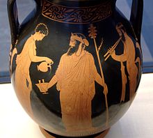 Peints sur un vase antique, dans un musée, en ocre sur fond noir, un homme avec une grande barbe noire, un sceptre et une couronne se fait verser un liquide dans une coupe par une femme ou un jeune homme. Un homme joue d'un instrument à vent derrière lui.