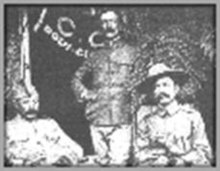 Photograph of three men at a Boy Scout event circa 1910. The man seated on the left is unidentified, Burnham is in the middle, standing, and Baden-Powell is on the right, seated. There is a table in front of the men. Baden-Powell is wearing his stetson hat, Burnham has no hat, and man on the left has a modest hat. Behind Burnham and to his right is a flag partially opened.