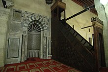 A niche made of multiple types of decorative stone is embedded in a wall, facing a red carpeted area. The niche is flanked by stone columns, and surmounted by a stone arch. The wall at the back of the niche is a semi-circular curve, with a geometric design covering most of it. The wall beside the niche also has patterned stone on it. To the right is a dark wooden structure, a narrow staircase with a lattice door at the foot of it, and lattice railings leading to a seat topped by a square wooden canopy.