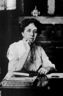 A white woman of middle age, seated, one hand on chin, wearing a white shirtwaist and glasses, with books on the table before her