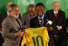 Frontale Farbfotografie von zwei Männern im Anzug, die gemeinsam ein brasilianisches Nationaltrikot mit der Aufschrift „Pelé 10“ halten. Pelé im Bild rechts hält einen Medaillenkasten.