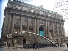 An entrance to the station in Bowling Green Park, with a glass canopy above it. Behind the entrance is the Alexander Hamilton U.S. Custom House.