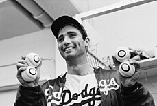 A baseball player in uniform and a jacket with "Dodgers" on it poses with four baseballs with zeroes on them, two in each hand, and smiles as he looks ahead.