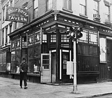 On the corner of a block is a building with large glass fronts on both sides; a sign displaying the tavern's name shines brightly above in red neon.