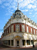 Photograph of a corner building with a curved front topped with a spire and flag pole. There are two rows of circular windows in the attics.