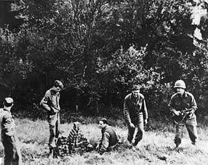 Four men in uniforms stand in a field looking at a pyramid shaped pile of cubes.