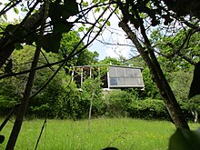 Photographie en couleurs en contre-plongée d’un bâtiment entouré de verdure. Des branches d’arbres au premier plan.