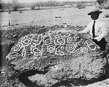 Photo taken in 1910 in Mexico of the very large Esperanza Stone, with Burnham standing to the right of the stone. The many inscriptions on the stone are sort of circular and have been filled in with white flour so they can be easily photographed. In the background is a desert landscape.