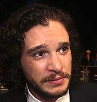 Kit Harington steht auf einem roten Teppich. Im Hintergrund ist ein Absperrzaun zu sehen, an dem einige Personen stehen. Harington trägt ein schwarzes Jackett, ein weißes Hemd, seine schwarzen, leicht gelockten Haare bis zum Hals sowie einen Kinnbart mit einem bis zu den Mundwinkeln reichenden Schnurrbart.