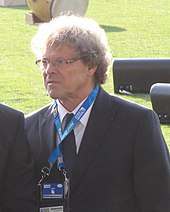 Un homme en costume porte un badge autour du cou et des lunettes sur le nez. Il a des cheveux frisés gris.