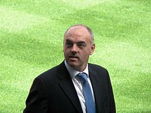 A balding man with a moustache in a suit, looking up, standing on grass.