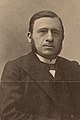 Photographie noir et blanc d'un homme en buste costumé avec un trois pièces au nœud papillon et portant des rouflaquettes.