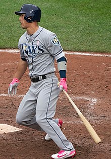 A man in a gray baseball uniform that says "Rays", a navy batting helmet, and pink batting gloves stands in the batters box after swinging at a pitch