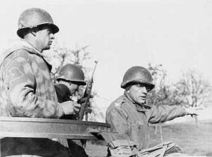 Three men in uniforms with steel helmets. One holds a rifle; another, wearing glasses, points to something in the distance.