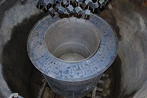 A circular hole, within which is another cylinder made of grey bricks. Black cubes are suspended from strings above.