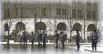 photo d'une avenue bordée d'immeubles imposants. L'avenue est coupée par une rangée de soldats en armes.