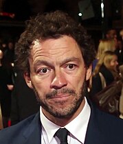 Dominic West steht auf einem roten Teppich. Im Hintergrund ist ein Absperrzaun zu sehen, an dem einige Personen stehen. West trägt ein dunkelblaues Jackett, ein weißes Hemd, eine schwarze Krawatte, seine braunen, leicht gelockten und abstehenden Haare bis zu den oberen Ohren sowie einen dünnen, struppigen, bis zu den Ohren reichenden Backenbart mit einem bis zu den Mundwinkeln reichenden Schnurrbart.