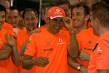 A group of people dressed in orange celebrate, with Hamilton in the middle wearing a baseball cap.