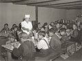 Une trentaine de garçons et filles du niveau primaire sont assis sur des bancs à de longues tables rectangulaires et mangent, qui dans un bol, qui dans une assiette, surveillés par une femme en uniforme et coiffe blanche, le repas servi grâce aux surplus alimentaires.