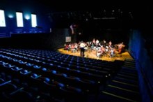 Empty blue seats and a small orchestra on a lighted stage, as seen from the rear of an auditorium