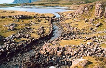 A stone lined ditch of primitive construction leads from a small lake. Rocky heathland lies on either side and there are tall cliffs in the distance.