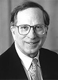 A man wearing glasses with a tie and a suit jacket looks to the camera and smiles