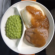 Photographie d’une assiette en plastique moulé à deux compartiments ; dans le plus petit, une matière verte ressemblant à des petits pois en sauce, dans le grand une plus grosse boule d’aliment ressemblant à de la purée de pomme de terre ou de riz, nappée partiellement d’une sauce gluante brune qui recouvre aussi la boule de nourriture contigüe, ressemblant à une purée de foie.