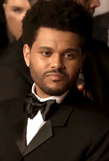 Tesfaye (the Weeknd), wearing a black suit with a bowtie, and with a neutral expression on his face, averts his gaze from the camera.