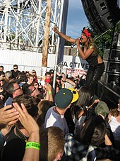 A dark-skinned woman wearing a black hat and black clothing holding a microphone while performing on stage to a crowd of people. The performance is taking place outdoors and part of a rollercoaster is visible in the background.