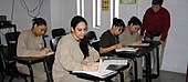 Quatre détenues en uniforme beige sont assises à des bureaux. Un professeur surveille l'une d'entre elles.