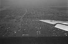 Black and white aerial image of a city, with virtually all streets in the photo flooded. The left wing of the aircraft from which this photo was taken is also visible at center-right.