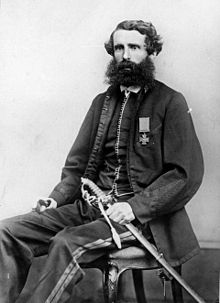 A black and white photograph of a bearded man in a jacket seated on a chair and holding a sword below the hilt