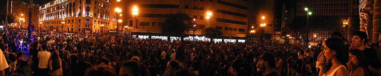 A huge crowd of people is gathered in a crowded public street at night in São Paulo outside the Municipal Theater