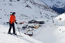 Photographie d'un skieur devant le centre d'une station de ski, avec les chalets, les remontées mécaniques et les skieurs en arrière-plan, le tout au pied des montagnes très enneigées.