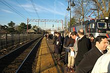 Mostly men wait at a train station with an empty track to their left and a train and leafless deciduous trees behind them.