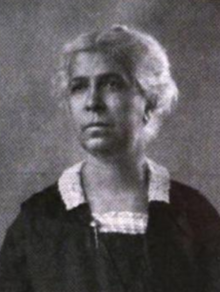 An older Black woman with grey hair, wearing a black dress with white lace collar detail