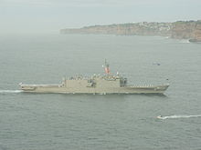 A large grey warship underway. Flags are being flown from the ship's mast, and white-uniformed people line the decks. Brown cliffs line the horizon for the right two-thirds of the image