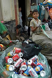 Devant une maison, un enfant ganté tient la lanière plastique d’un sac fermé, quatre à cinq fois pour gros que le garçonnet. Tout autour, ce ne sont qu’autres sacs plastiques, de toutes dimensions, ouverts ou éventrés, contenant tous des canettes, boîtes de conserve ou bouteilles usagées. Dans l’encoignure de la porte, près d’une chaise vide, une femme en robe à carreaux bleus et blanc regarde discrètement le photographe comme le fait aussi une autre, chapeautée, qui est partiellement dissimulée par l’enfant.