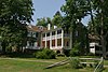 A two story house with dormers and a porch, with trees around it in the sunshine