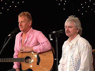 Two grey-haired men sitting at microphones, one holding a guitar