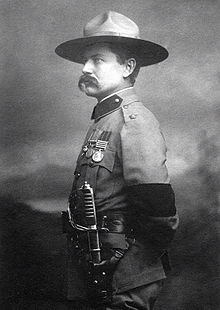 Photo of Burnham taken in 1901 in London after his investiture with the cross of the Distinguished Service Order by King Edward VII. He is dressed in British Army uniform and standing at attention, facing right. On his left arm is a black armband worn in mourning for the recent death of Queen Victoria. He is wearing his stetson hat and a sword on his left side. He sports a large handlebar mustache.