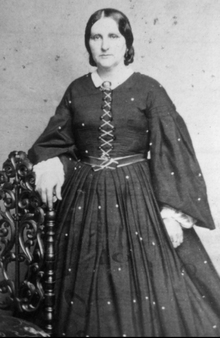 19th-century B&W portrait photo of a standing woman wearing a dark long dress with a white collar.