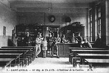 Photographie de la cantine militaire de Saint-Avold : de part et d’autre d’une allée centrale, des tables avec bancs sont disposées. Au bout de l’allée, un comptoir servant de bar auquel sont adossés des soldats et une fillette posant pour le photographe.