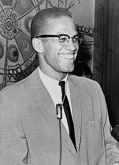 A 38-year-old man in a suit and tie is grinning. He wears glasses and has a microphone around his neck